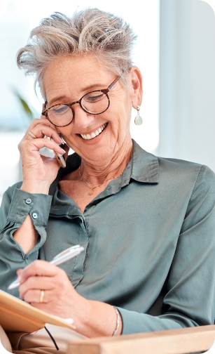 Nundah General and Emergency Dental Older woman smiling while talking on the phone and writing in a notebook.